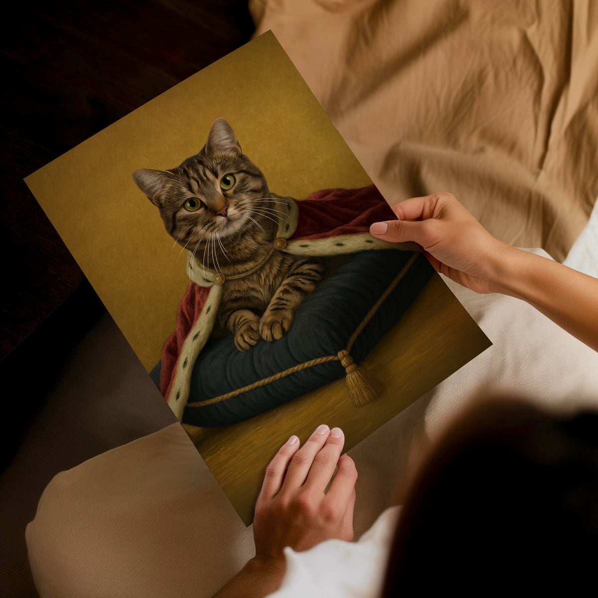 A classic style, golden backdrop wall art. A person holds a custom pet portrait print showing a tabby cat with a burgundy cape sat upon a plain pillow, dressed in medieval royal attire, set against a cozy bedroom background with beige bedding.