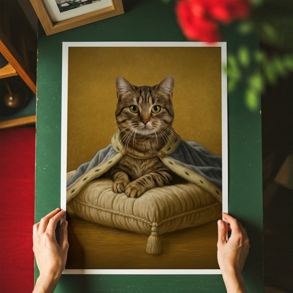 A classic golden backdrop framed-style art print showing a tabby cat in royal light blue attire seated on a subtle ivory cushion, held up by two hands over a green tabletop. The scene includes blurred red flowers and a wooden frame in the background.