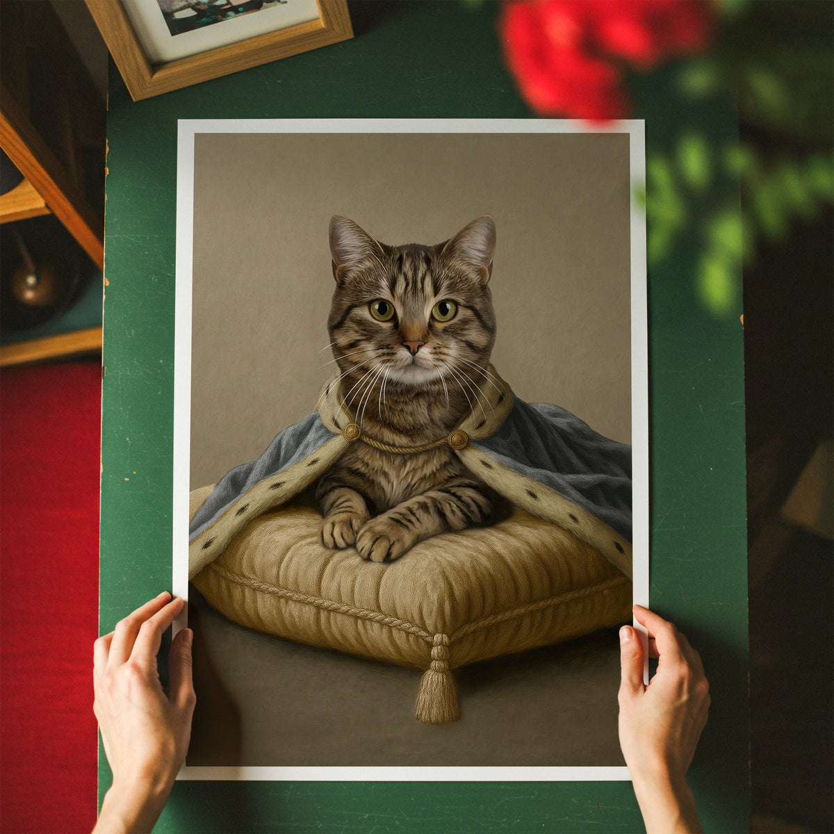 A modern natural beige backdrop framed-style art print showing a tabby cat in royal light blue attire seated on a subtle ivory cushion, held up by two hands over a green tabletop. The scene includes blurred red flowers and a wooden frame in the background.