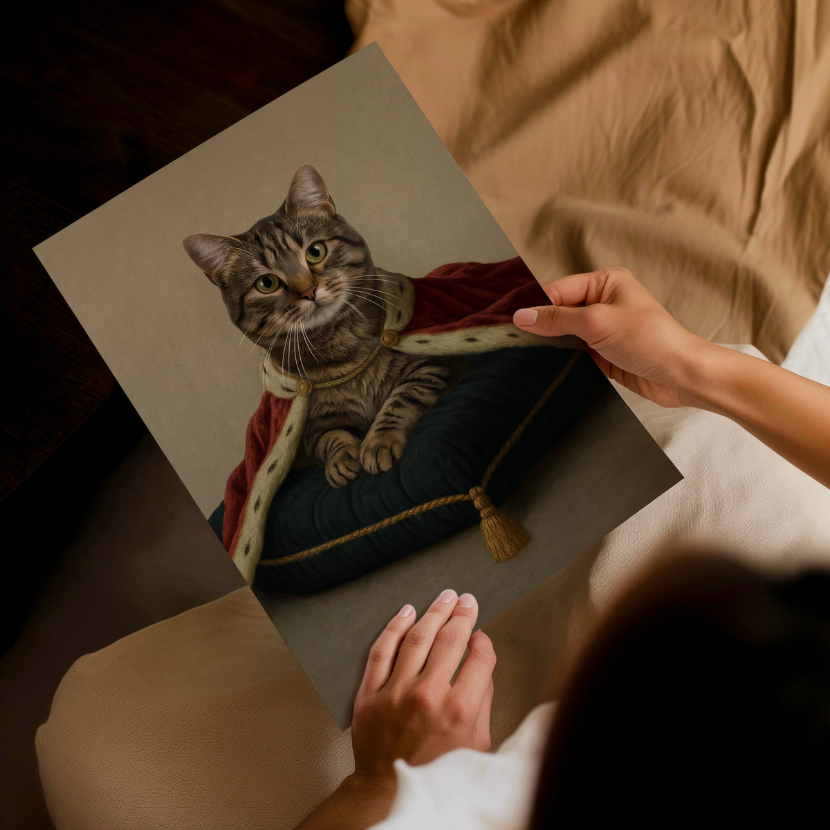 A modern style, beige concrete-like backdrop wall art. A person holds a custom pet portrait print showing a tabby cat with a burgundy cape sat upon a plain pillow, dressed in medieval royal attire, set against a cozy bedroom background with beige bedding.